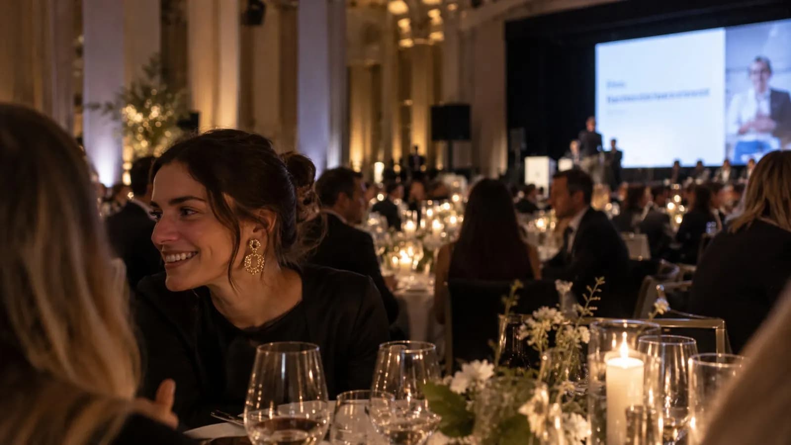 Guests at a candlelit gala dinner, a speaker on stage behind them